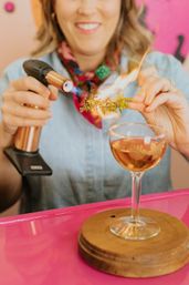 Person using a handheld culinary torch to ignite a rosemary sprig over a craft cocktail in a coupe glass on a wooden coaster at a bright pink bar counter