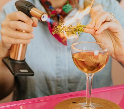 Person using a handheld culinary torch to ignite a rosemary sprig over a craft cocktail in a coupe glass on a wooden coaster at a bright pink bar counter