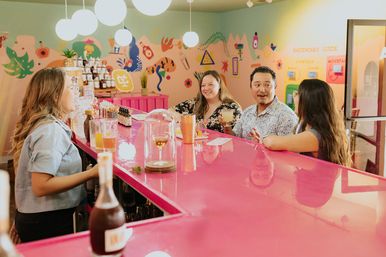 Colorful cocktail bar interior with a bright pink counter, hanging globe lights and a tropical wall mural; a group of four laughing and enjoying cocktails at the bar.