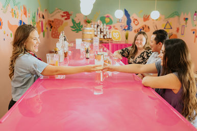 Group of friends sharing cocktails at a bright pink bar counter with a colorful mural wall in a cheerful indoor tasting room