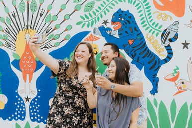 Three friends smiling and holding cocktails while taking a selfie in front of a colorful mural with a peacock, blue tiger, and leafy botanical motifs.