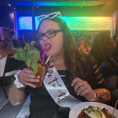 Person wearing a celebratory sash and rhinestone sunglasses enjoying an oversized Bloody Mary garnished with lettuce and bacon in a colorful nightclub or bar.