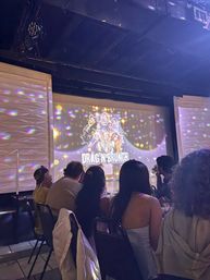 Indoor drag brunch show with seated audience silhouettes watching a colorful projected poster of drag performers on stage, disco light reflections on textured walls and lively brunch-theater atmosphere.