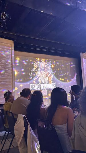 Indoor drag brunch show with seated audience silhouettes watching a colorful projected poster of drag performers on stage, disco light reflections on textured walls and lively brunch-theater atmosphere.