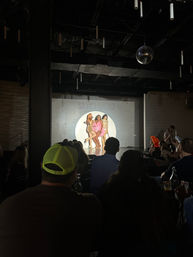 Live drag brunch show with three performers in a spotlight on a small nightclub stage, mirrored disco ball overhead and a seated audience watching