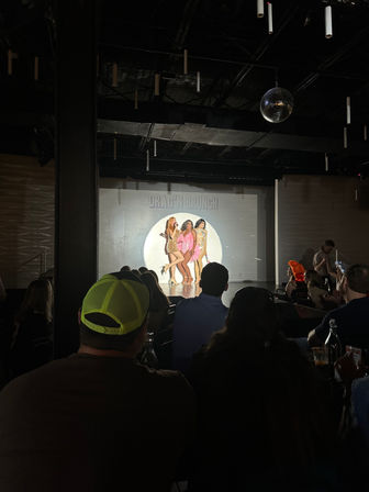 Live drag brunch show with three performers in a spotlight on a small nightclub stage, mirrored disco ball overhead and a seated audience watching
