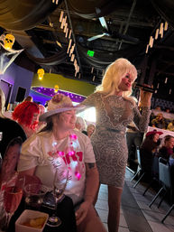 Drag performer in a sequined dress sings into a microphone while engaging a smiling guest in a cowboy hat at a lively indoor cabaret-style nightclub with skull lights and hanging candle decorations.