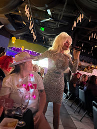 Sparkling drag performer in a rhinestone silver dress and blonde wig sings into a microphone while interacting with seated audience wearing festive hats at a lively indoor cabaret-style nightclub.