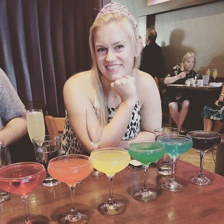 Smiling woman wearing a party tiara at a restaurant table lined with colorful rainbow cocktails in coupe glasses