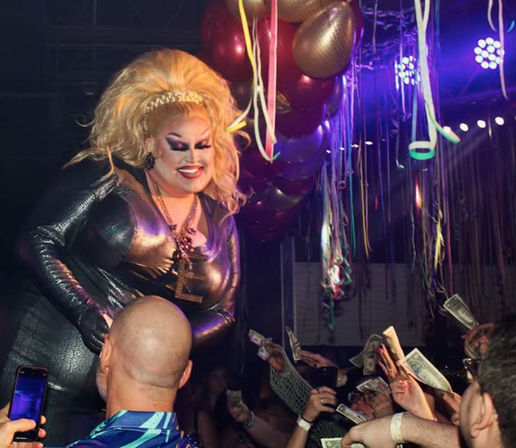 Drag show performer in a metallic dress and big blonde wig smiles on a nightclub stage as an enthusiastic crowd throws dollar bills amid balloons and colorful streamers.