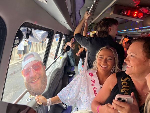 Smiling bride-to-be in a sequin dress and sash laughing with friends on a lively party bus, holding a large cardboard face cutout by the window with an urban street visible outside — bachelorette party vibe.