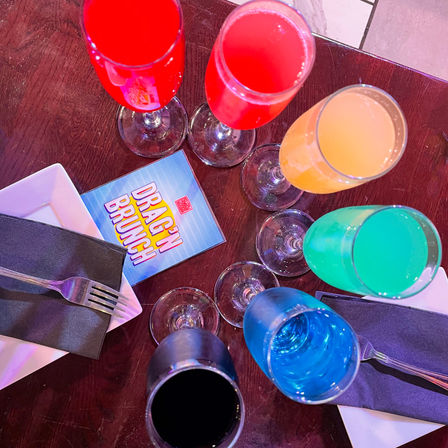 Overhead shot of six vibrant champagne-flute cocktails in rainbow colors surrounding a 'Drag'n Brunch' card on a dark wooden table, with plates, black napkins, and forks — playful brunch setting.