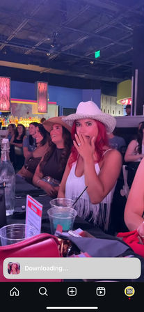 Surprised woman in a sparkly white cowboy hat showing an engagement ring during a girls' night at a colorful indoor bar with neon lighting, drinks and friends wearing hats