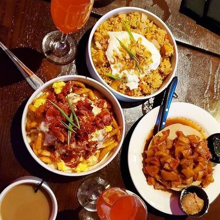 Brunch spread on dark wooden table: loaded breakfast fries with bacon and eggs, savory rice bowl with sour cream and chives, caramelized apple pancake, coffee and orange cocktails.