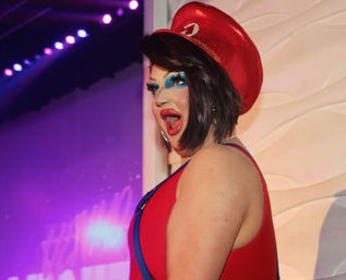 Drag performer in a red sailor-style hat and matching red outfit, wearing dramatic blue eye makeup and bright red lipstick, posing on a purple-lit nightclub/cabaret stage.