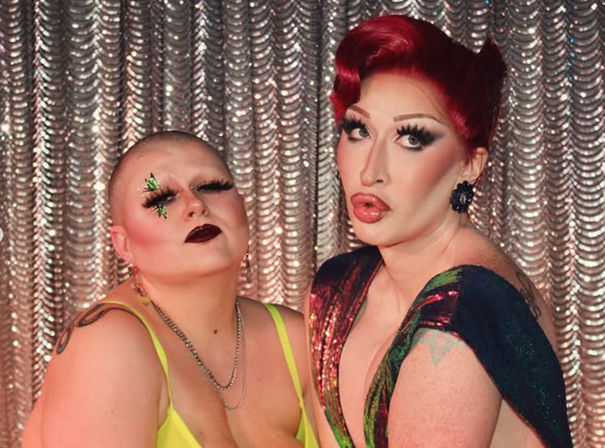 Two glamorous drag performers posing on a cabaret stage with a silver sequin curtain — bald performer in neon green and jeweled lashes, red‑haired performer in shimmery multicolored gown and vintage waves.