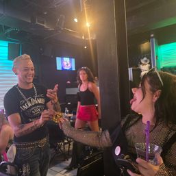 Playful nightlife scene in a neon-lit nightclub: a tattooed man laughs as he hands a gold-wrapped item to a woman sticking out her tongue while she holds a drink.