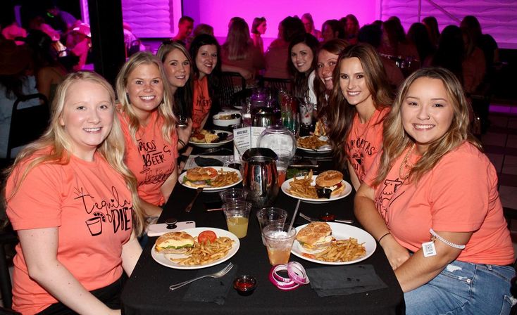 Cheerful group of women in matching coral T-shirts seated at a long restaurant table with burgers, fries and drinks under purple stage lighting.