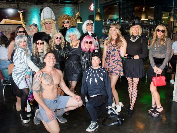 Lively group photo at a themed costume party in an indoor bar — colorful wigs, sunglasses, hats and eclectic outfits, one shirtless man kneeling in front, pendant lights and a stocked bar in the background.