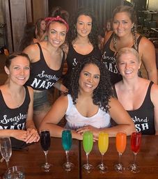 Six women in matching tank tops smiling and posing at a wooden bar table with a row of colorful cocktails in champagne flutes — festive, fun group photo in a lively bar setting.