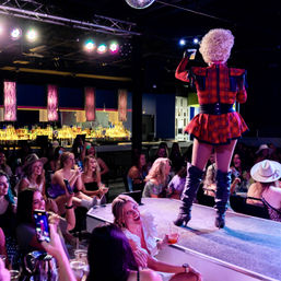 Drag performer in red plaid costume and thigh-high boots on a nightclub stage, lively cheering audience with drinks and phones and an illuminated bar backdrop