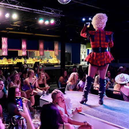 Drag performer in red plaid costume and thigh-high boots on a nightclub stage, lively cheering audience with drinks and phones and an illuminated bar backdrop
