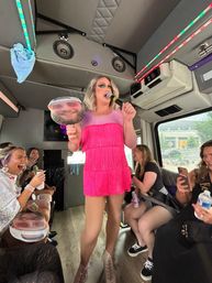 Drag performer in bright pink fringe dress holding a microphone and a face cutout, entertaining laughing passengers inside an LED-lit party bus.