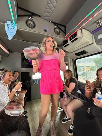 Drag performer in bright pink fringe dress holding a microphone and a face cutout, entertaining laughing passengers inside an LED-lit party bus.