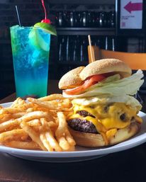 Diner-style double cheeseburger with melted cheddar, iceberg lettuce and tomato on a toasted bun, served with crispy French fries and a vibrant blue lime cocktail garnished with cherry and lime.