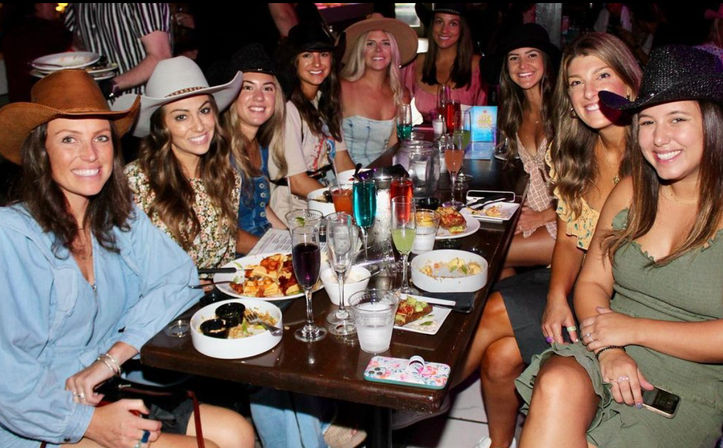 Smiling group of women in cowboy hats enjoying a lively restaurant dinner with colorful cocktails and shared plates — casual night out with friends.