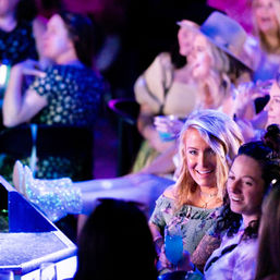 Smiling women in a colorful nightclub audience watching a live performance under purple-blue lights; a blonde woman holds a bright blue cocktail while friends laugh and sparkly boots glimmer near the stage.
