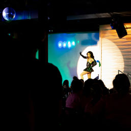Drag performer in a green sequin mini-dress and long glove strikes a pose under a spotlight on a nightclub stage with colorful projection lights and a disco ball, silhouetted audience watching.