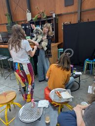 Smiling woman holding a fluffy dog in an industrial-chic indoor cafe, friends chatting by a wood-paneled counter while another person works on a laptop; checkered pizza plates and colorful stools in foreground.