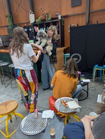 Smiling woman holding a fluffy dog in an industrial-chic indoor cafe, friends chatting by a wood-paneled counter while another person works on a laptop; checkered pizza plates and colorful stools in foreground.