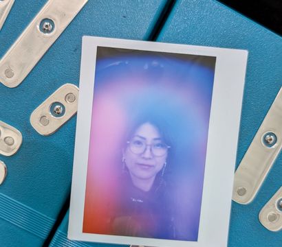 Polaroid-style instant photo of a woman wearing glasses surrounded by a pink-purple-blue aura, laid on a teal metal equipment case with silver hardware.