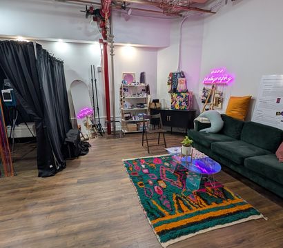 Bright, cozy indoor photo studio with a black-curtained photo booth and rainbow bead curtain, full-length mirror, props shelf, neon pink sign, green velvet sofa with colorful pillows, vibrant boho rug and acrylic coffee table on wood floors.