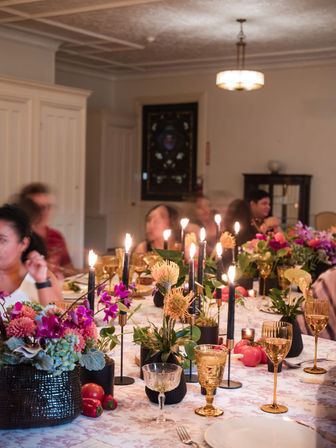 Warm candlelit dinner table in an elegant dining room with tall black tapers, gold goblets, mixed pink and purple floral centerpieces, decorative fruit and glassware, and blurred guests chatting in the background