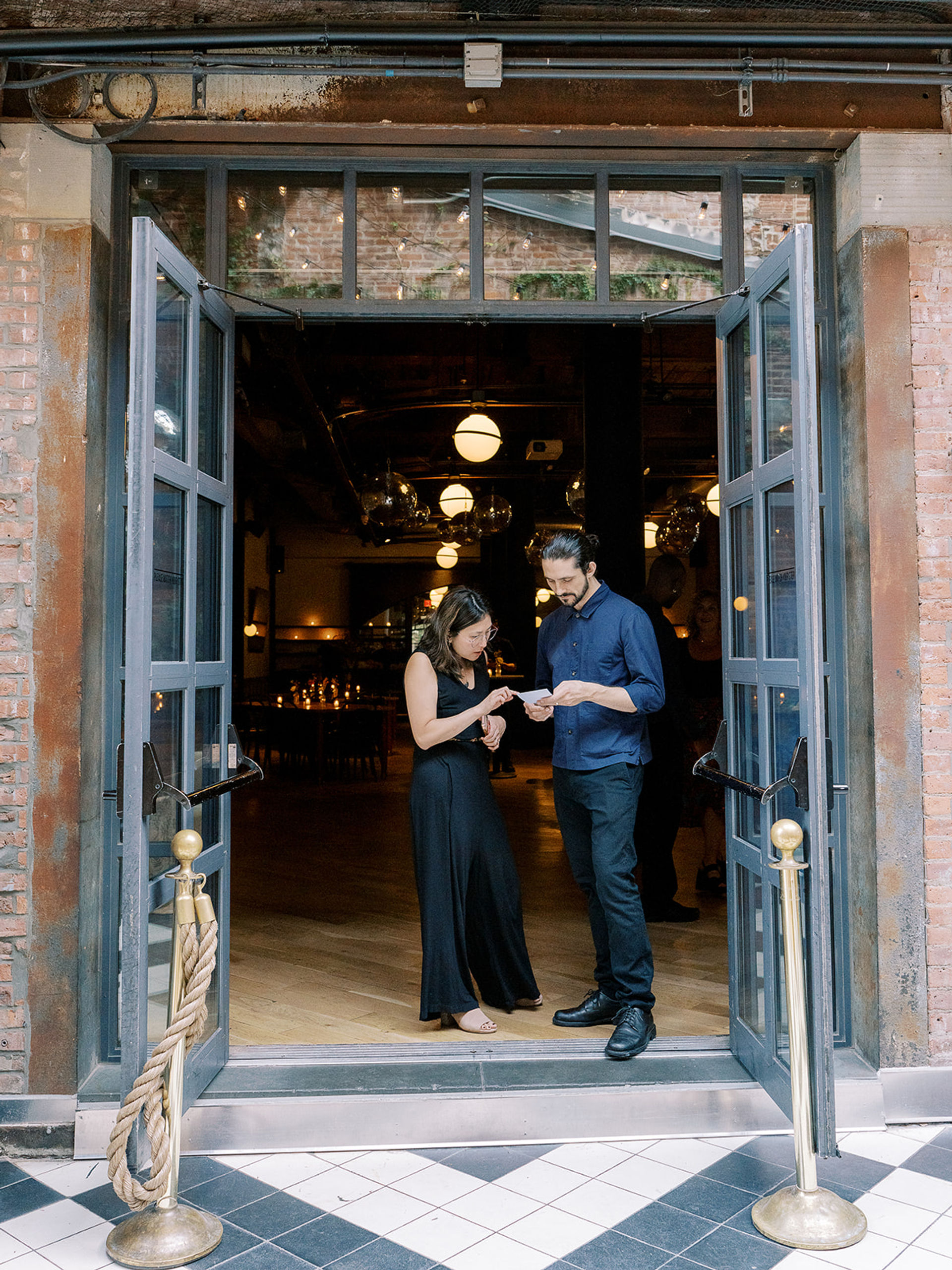 Two people checking a reservation at an open glass-door urban restaurant entrance with brass stanchions, rope barrier, tiled entry and warm hanging globe lights inside.