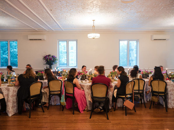 Candlelit dinner party in a bright dining room with an ornate ceiling, long floral-adorned table, guests seated by city-view windows on a wooden floor.