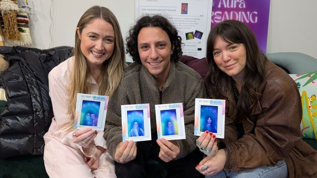 Three smiling adults seated on a couch at an indoor wellness pop-up holding four colorful aura-style instant photos showing blue and purple energy halos