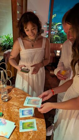 Friends in white outfits at a cozy, dimly lit restaurant table flipping colorful aura-style photo cards on a marble top, with cocktails, a water bottle, plants and string lights in the background.