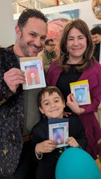 Smiling family of three at an art gallery event holding colorful "See Your True Colors" aura photos, young boy with teal balloon in front of abstract paintings.