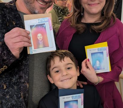 Smiling family of three at an art gallery event holding colorful "See Your True Colors" aura photos, young boy with teal balloon in front of abstract paintings.