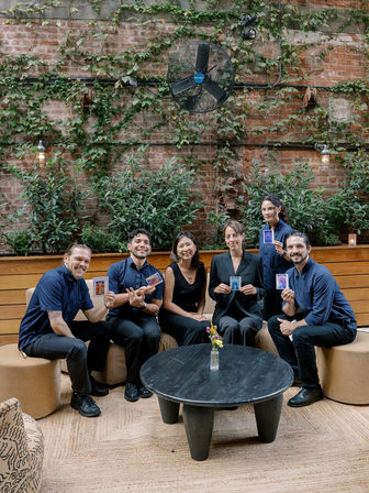 Six-person team portrait on a cozy urban courtyard patio with a brick wall draped in climbing ivy and a large wall fan, seated around a black round table with a small vase, each holding colorful instant photos.