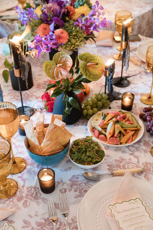 Candlelit dining table with colorful floral centerpiece and anthurium leaves, gold goblets, bowl of crackers, fresh fruit and salad on a patterned tablecloth — elegant intimate dinner setting.