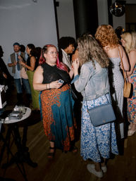 Indoor urban social gathering with people chatting under a disco ball — woman with pink hair and a colorful skirt holding a phone, talking to a friend in a denim jacket and blue floral dress.