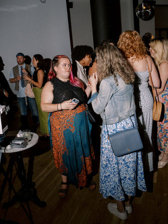Indoor urban social gathering with people chatting under a disco ball — woman with pink hair and a colorful skirt holding a phone, talking to a friend in a denim jacket and blue floral dress.