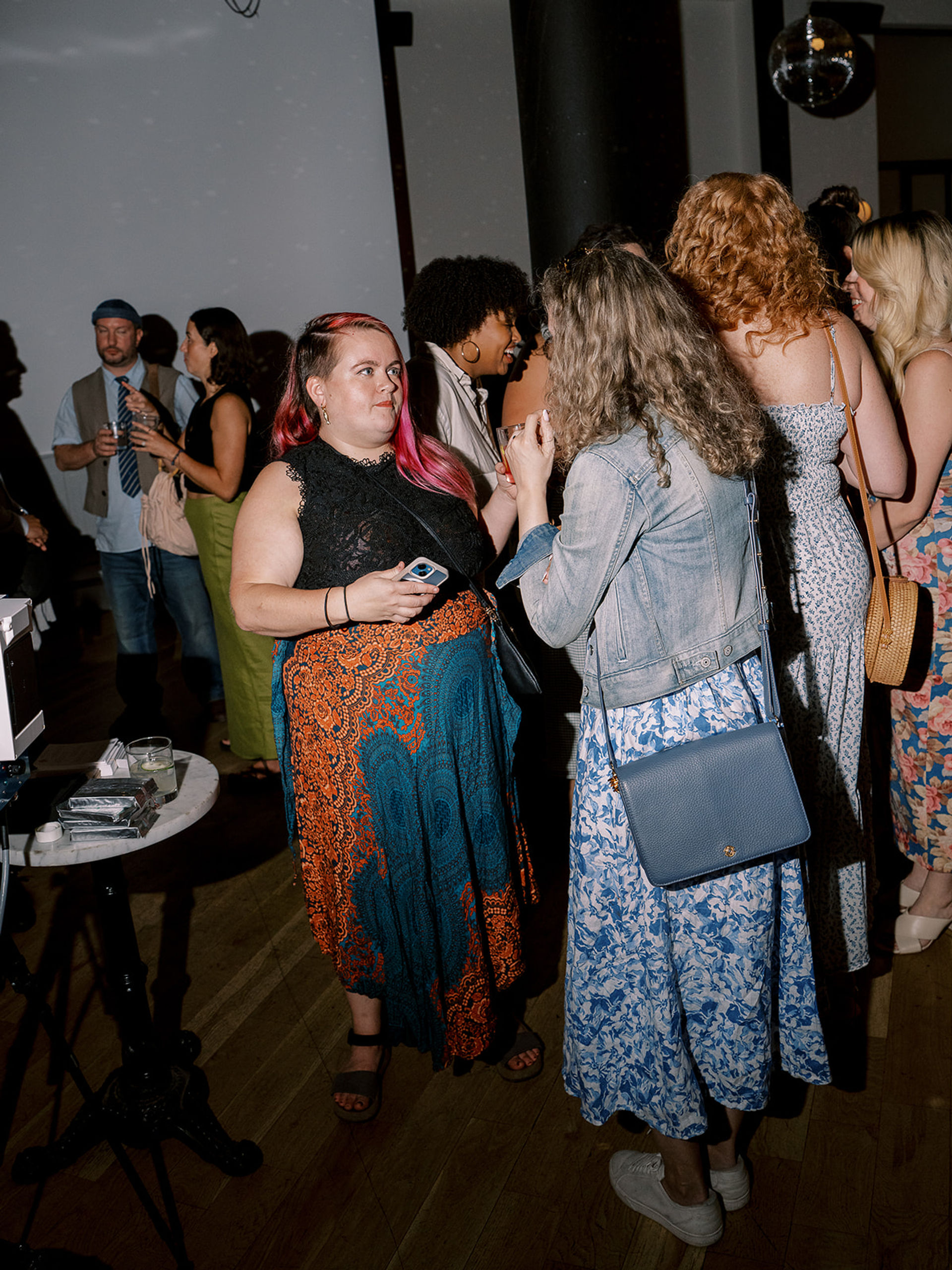 Indoor urban social gathering with people chatting under a disco ball — woman with pink hair and a colorful skirt holding a phone, talking to a friend in a denim jacket and blue floral dress.