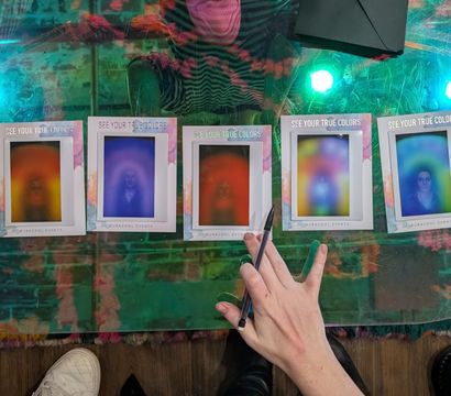 Seven colorful aura photo prints with rainbow halos lined up on a glass table at an indoor event, a hand holding a pen pointing at one print while multicolored lights reflect and shoes peek at the table edge.