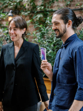 Two smiling professionals in an urban courtyard with a brick wall and greenery; a man in a navy shirt holds a small colorful photo card while a woman in a pinstripe blazer laughs.